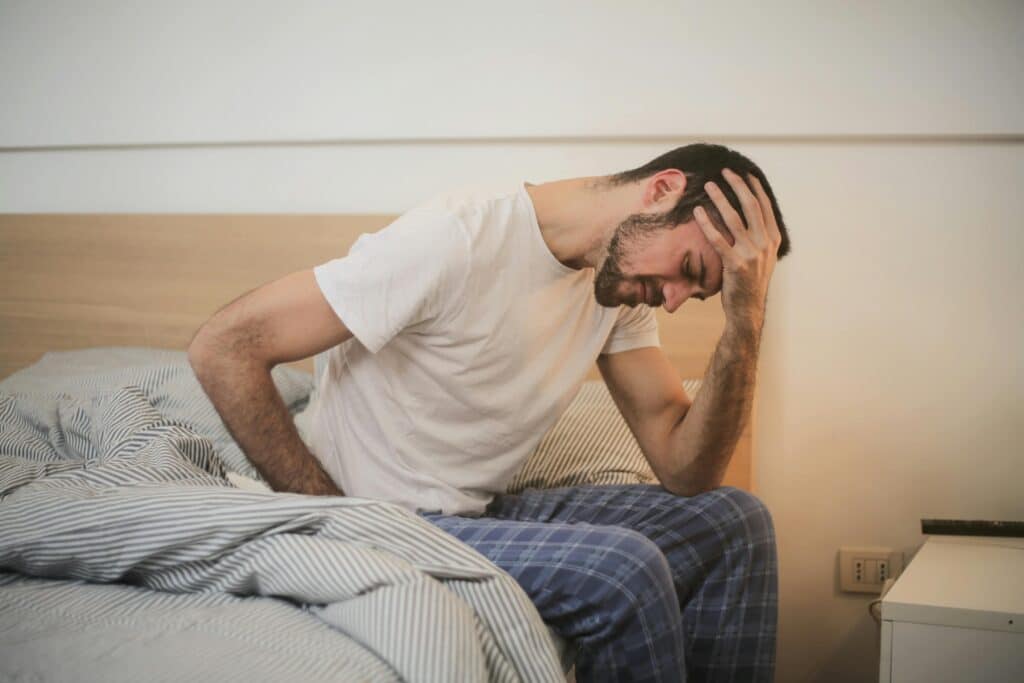 A young man worried about being laid off and having lay offs in his company in pajamas holding his head, sitting on a bed, appears to be experiencing a headache.
