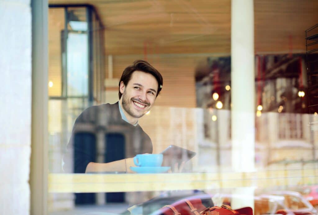 A cheerful man with a beard enjoys coffee at a café, viewed through a window.