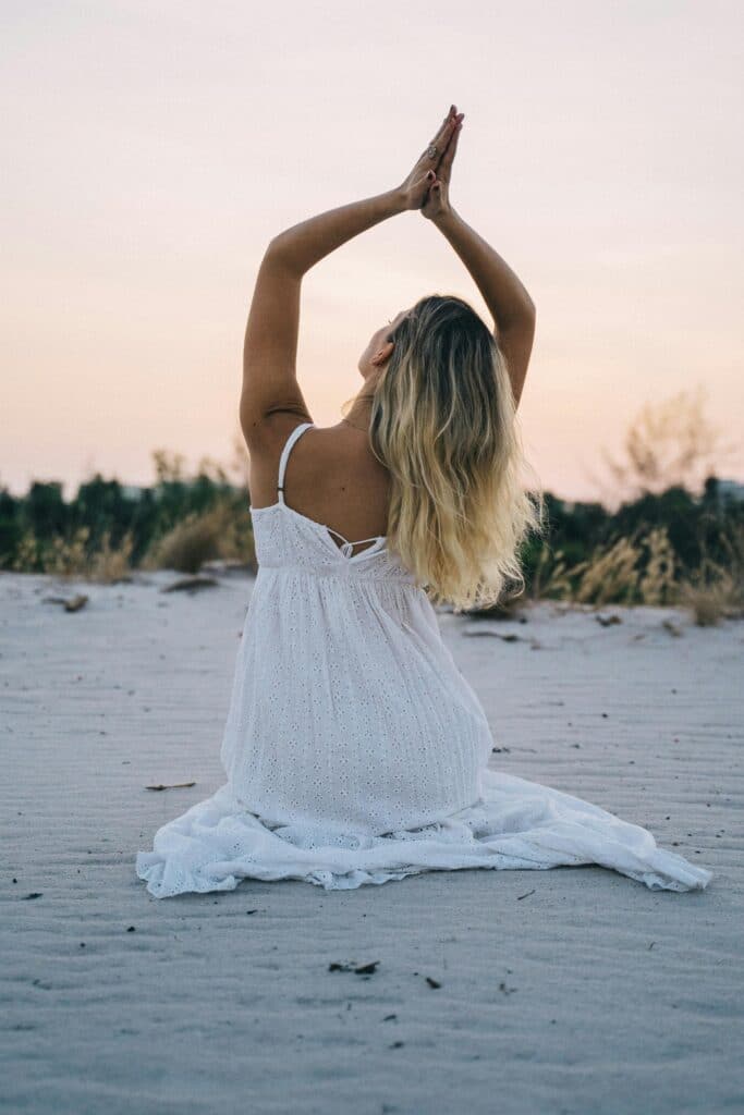 Woman in a white dress meditating on a serene beach during sunset, enhancing calmness and mindfulness.