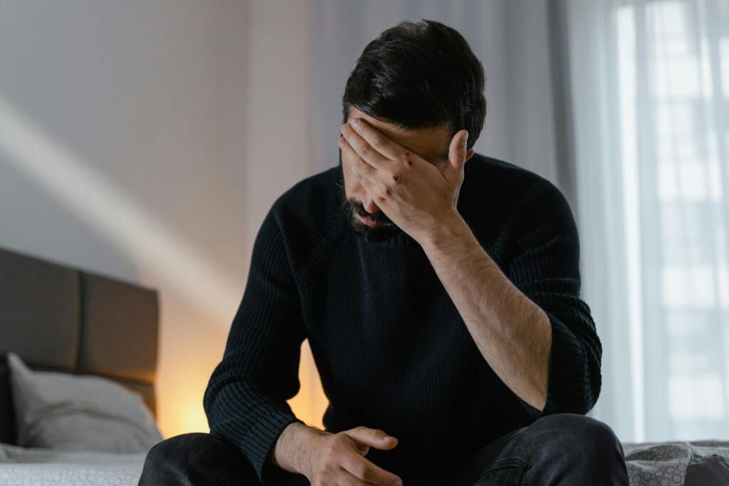 A man in distress sits on a bed indoors, covering his face with his hand.