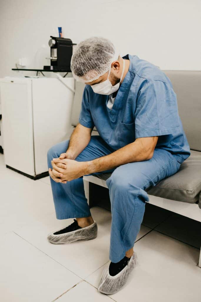 A male doctor in hospital attire sitting pensively, representing healthcare challenges.