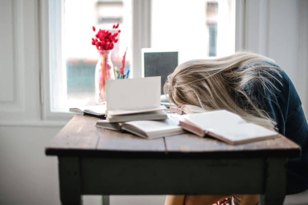 Blonde woman slumped over desk filled with books, showcasing study fatigue indoors symbolizing burnout and the need to learn how to be yourself
