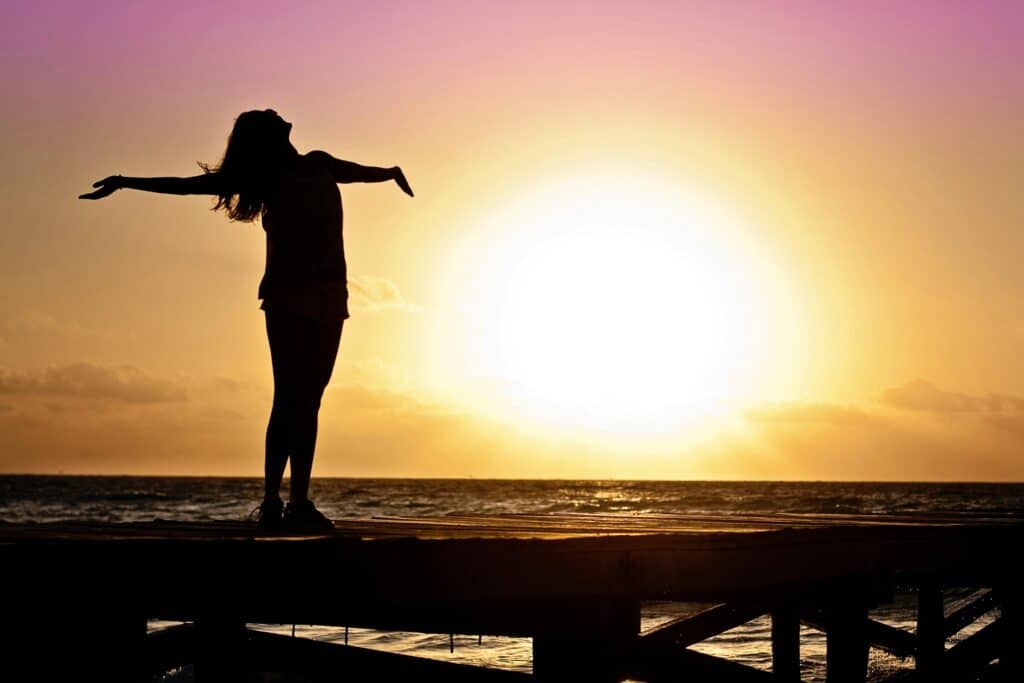 How to be yourself theme symbolized by A joyful silhouette of a woman standing on a dock at sunset, embracing the freedom of nature.