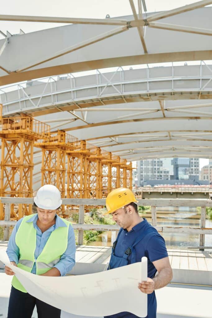 Two engineers on a construction site examining blueprints with steel structures in the background showing decision pressure and responsibility in mining, oil & gas and trades.”