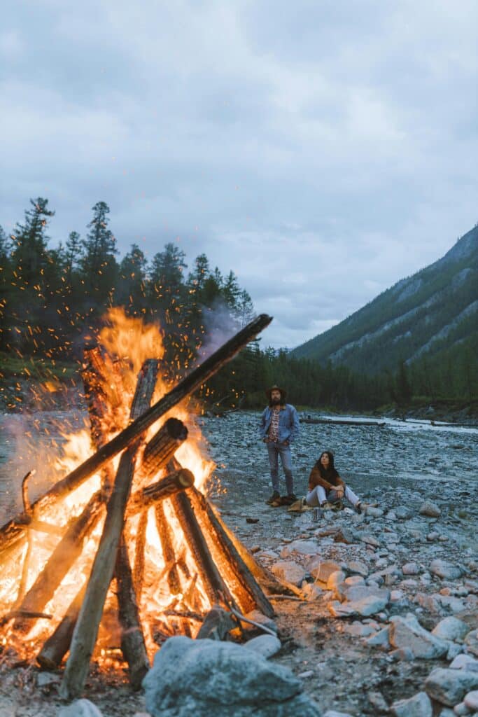 A couple sits by a large bonfire on a rocky riverside with mountains in the background.
