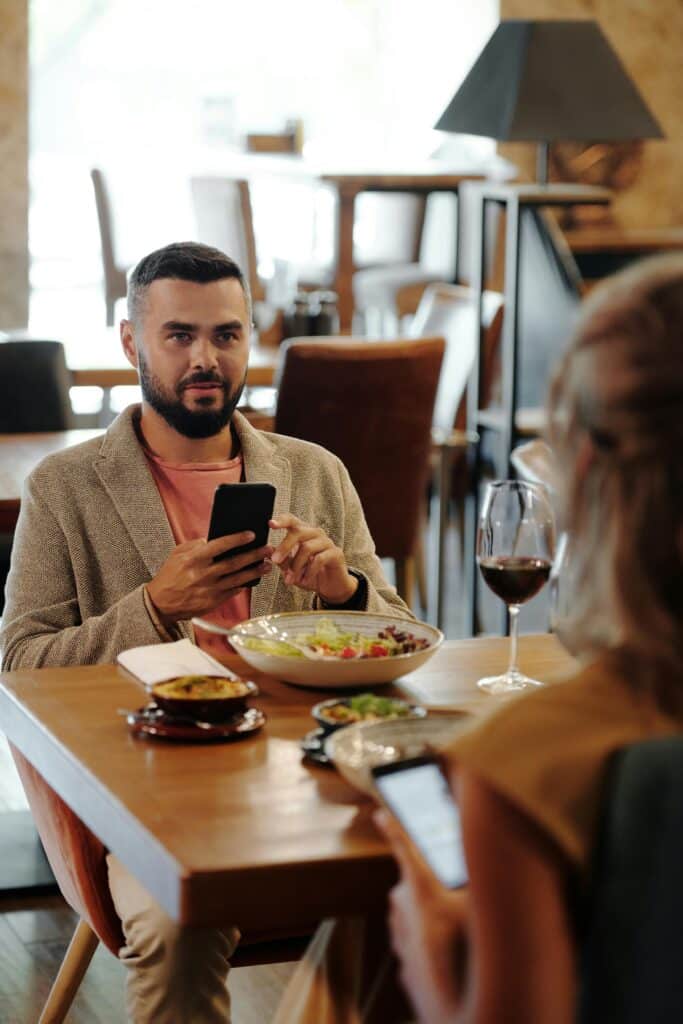 A man and woman sit at a restaurant table, using smartphones while dining.