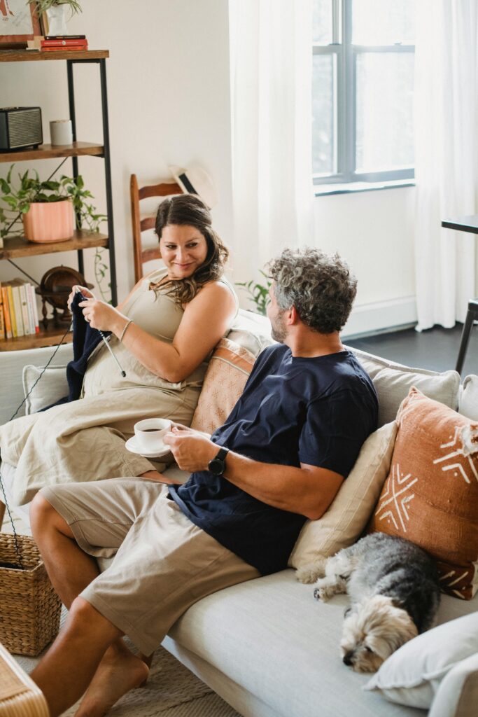 Cheerful pregnant female in dress knitting while sitting on sofa with bearded husband drinking coffee and sleeping dog