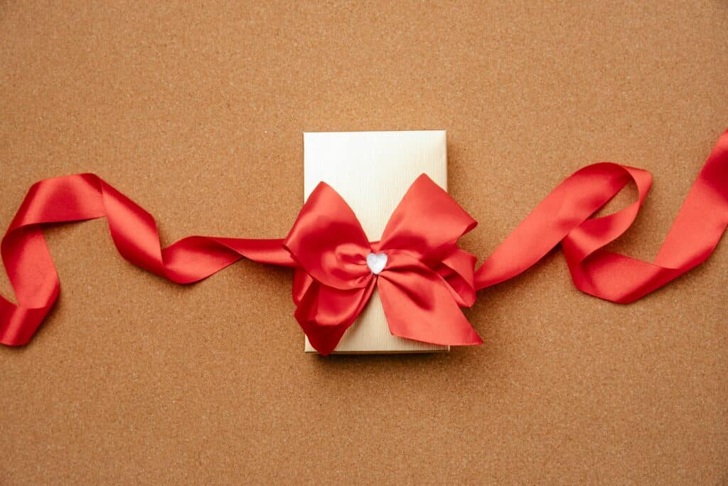 A beautifully wrapped gift box with a red satin bow and heart embellishment on a cork background.