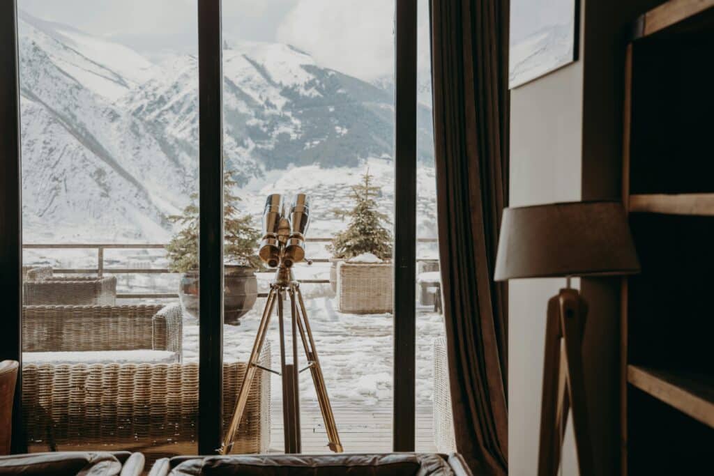 A cozy room with a telescope facing snow-covered mountains through large windows.