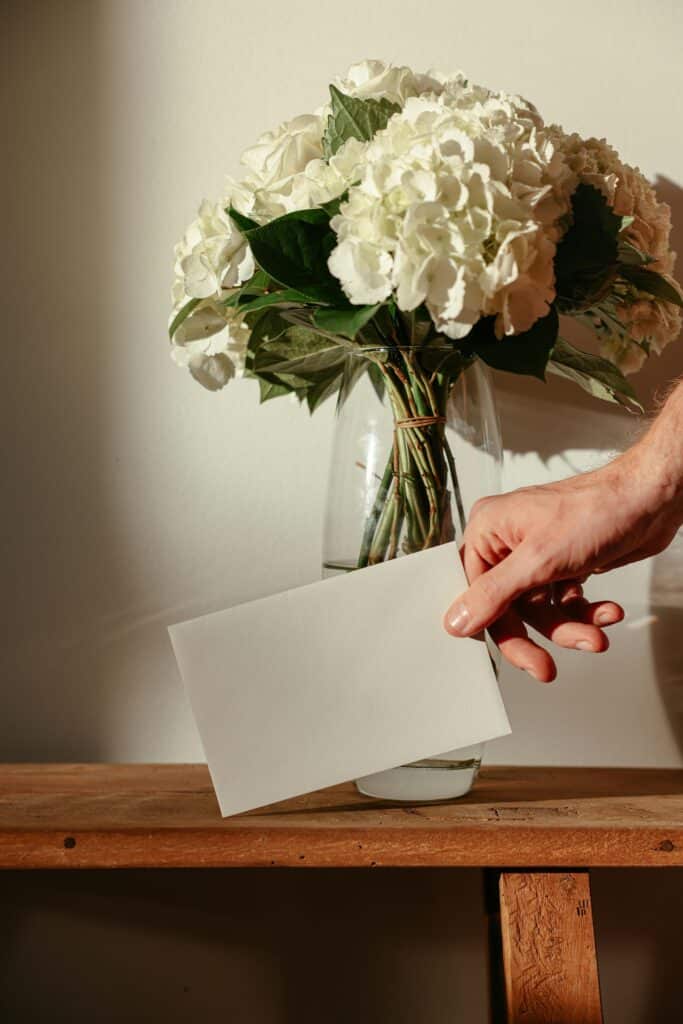 A hand holding a card next to a vase of white flowers on a wooden table.