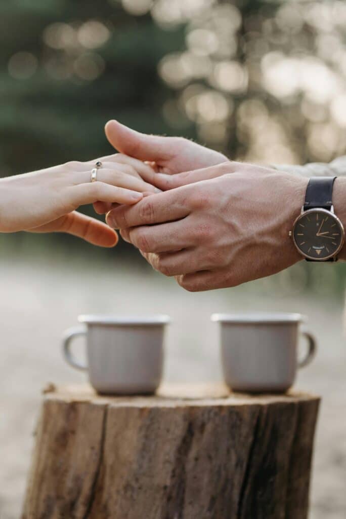 Close-up of a couple holding hands over a wooden stump with mugs outdoors, showcasing love and connection.