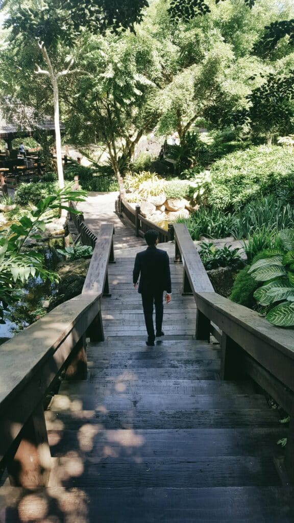 Person walking down stairs in a lush outdoor garden with tropical plants and wooden pathways.