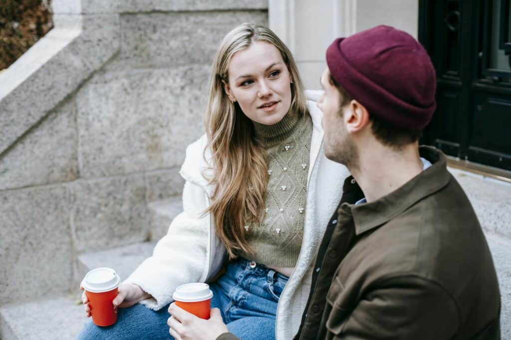 Man and woman talking sitting on steps outside house and having hot beverage in paper cups