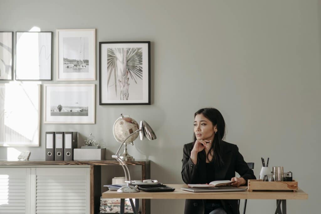 Asian woman in black blazer sitting at a desk in stylish home office, deep in thought.